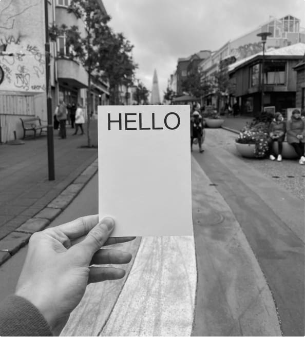 A white postcard with the word "hello" printed prominently in black held in front of a bustling European street.