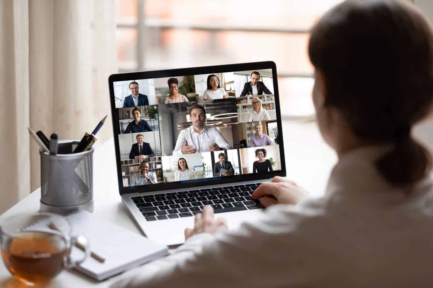 View over business lady shoulder seated at workplace desk look at computer screen where collage of many diverse people involved at video conference negotiations activity, modern app tech usage concept