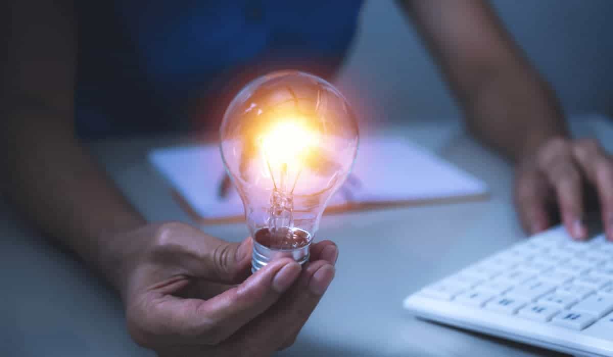 a hand holding a lit lightbulb above a desk with a keyboard and notebook on top