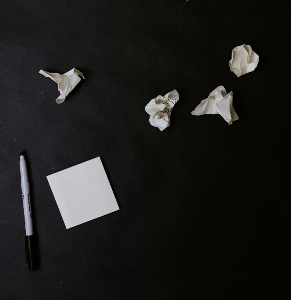 blank square notepad next to a pen with crumbled notes next to it on a black surface