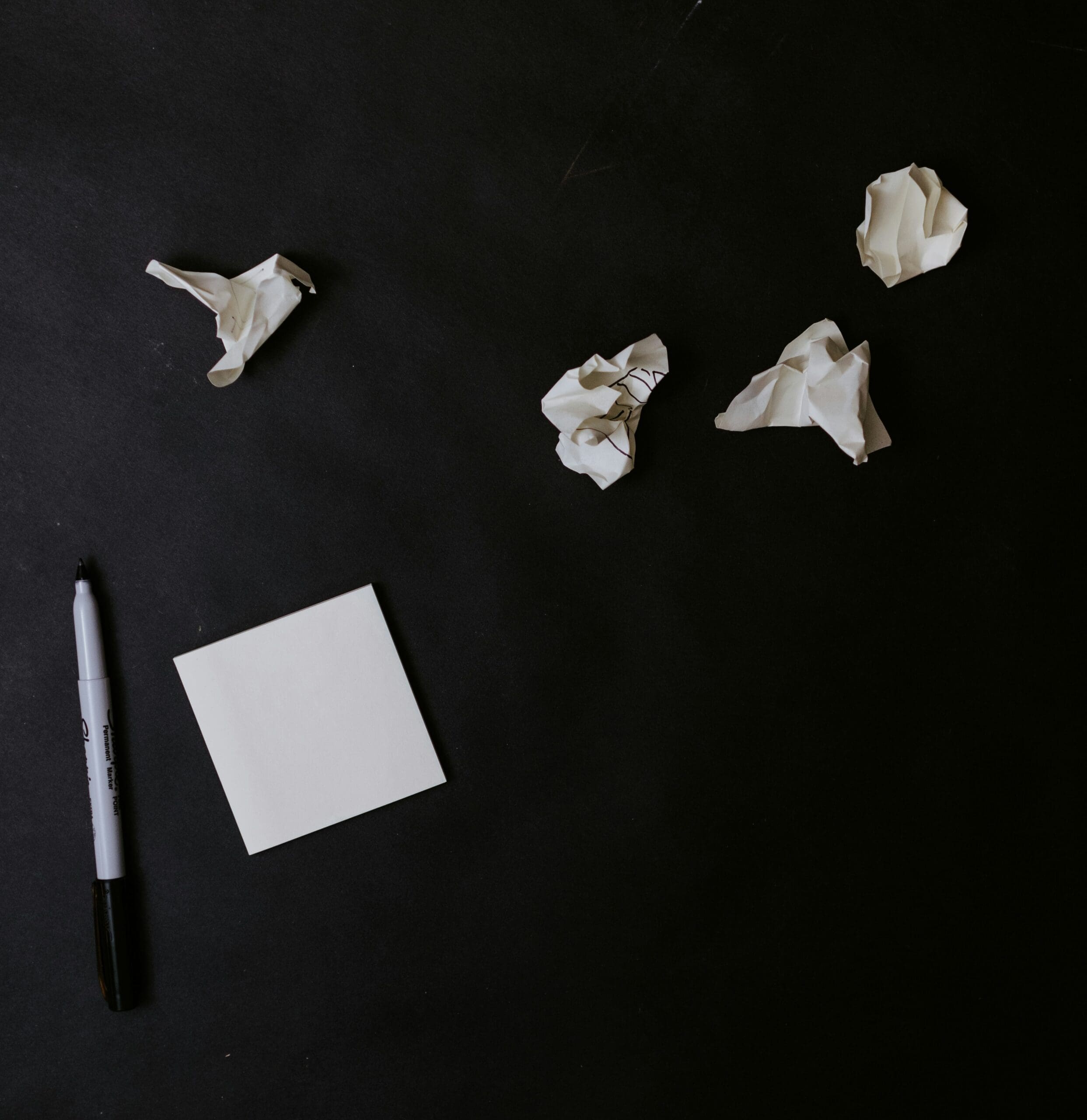 blank square notepad next to a pen with crumbled notes next to it on a black surface